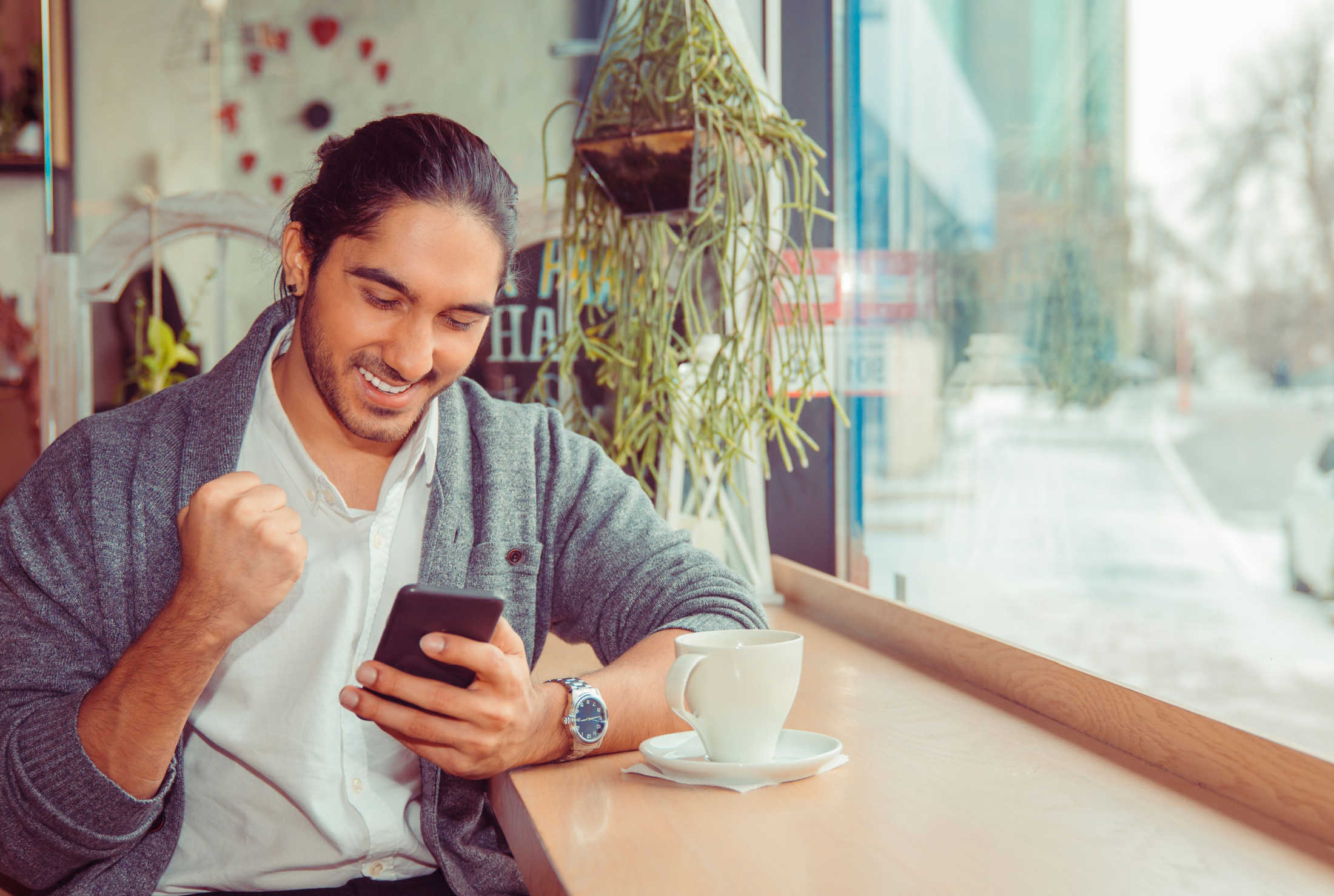 Happy young male professional using his smartphone while enjoying a coffee at a cafe bar.