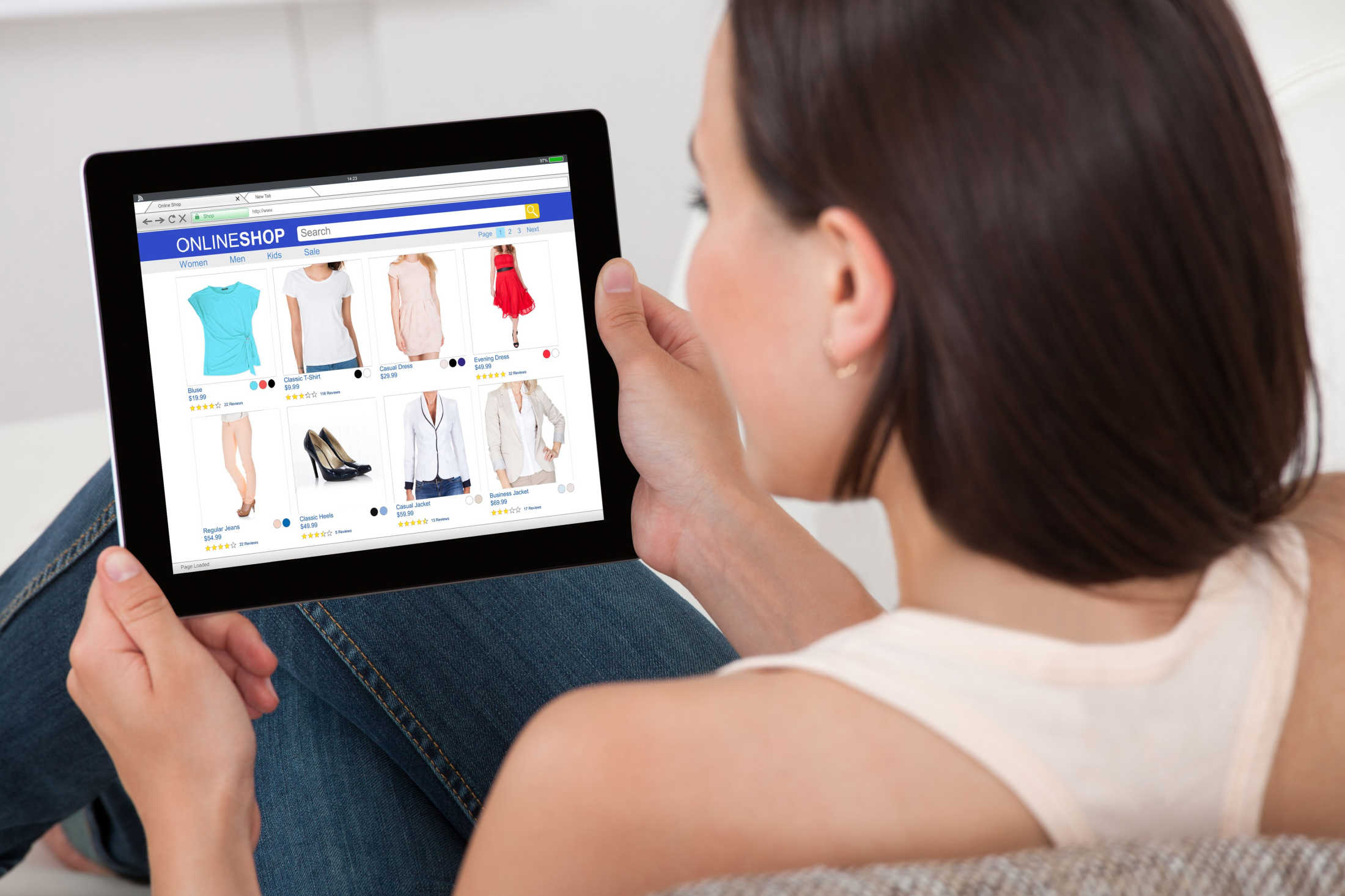 Over the shoulder view of a woman sitting on her couch while online shopping for female clothing on a tablet.